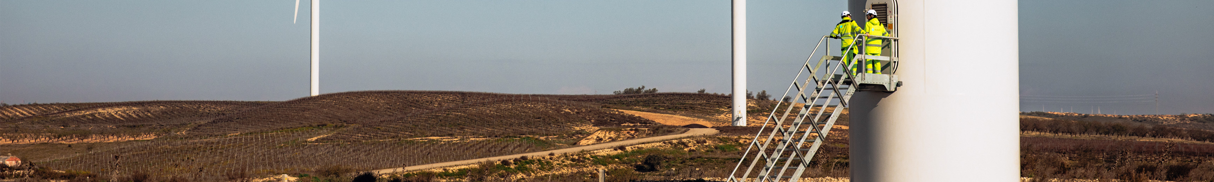 Nærbilde av masten til en vindturbin. To ansatte på en trapp. To vindturbiner i bakgrunnen.